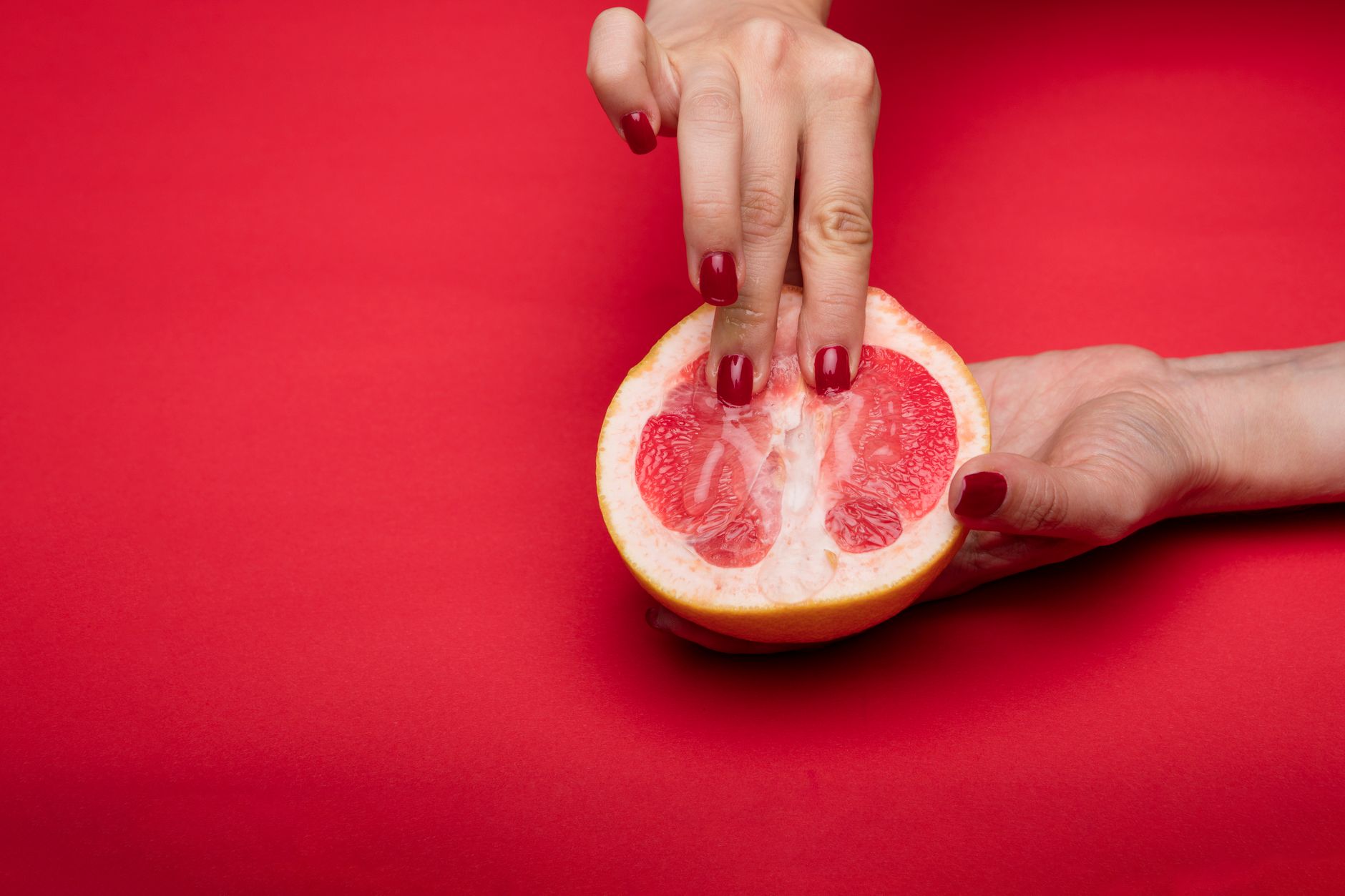 Close-up of hands with red nails holding a halved grapefruit on a red background, conceptual and vibrant.