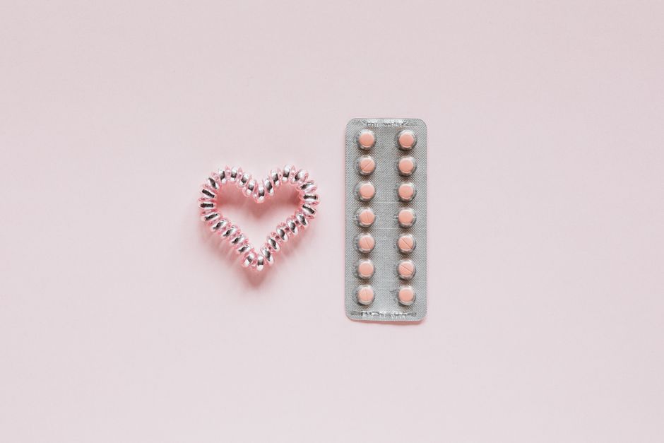 From above of whole blister of pills and shiny handmade heart of coil hair tie on pink surface