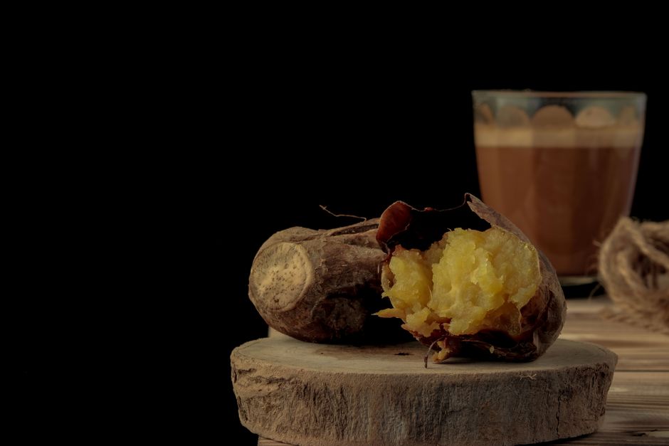 Still life of roasted sweet potato with a glass of coffee on a wooden table against a dark background.