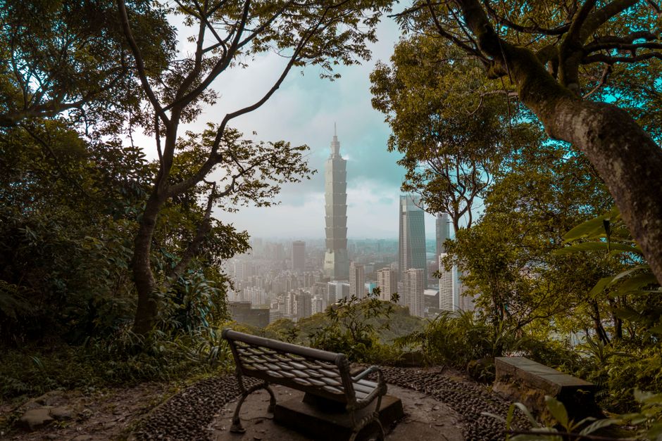 A picturesque view of Taipei 101 skyscraper framed by lush greenery and a park bench.