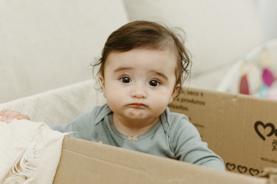Cute baby sitting in a cardboard box with a confused expression, indoors.